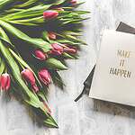 A notebook with 'Make It Happen' inscription beside a bouquet of pink tulips, capturing inspiration.