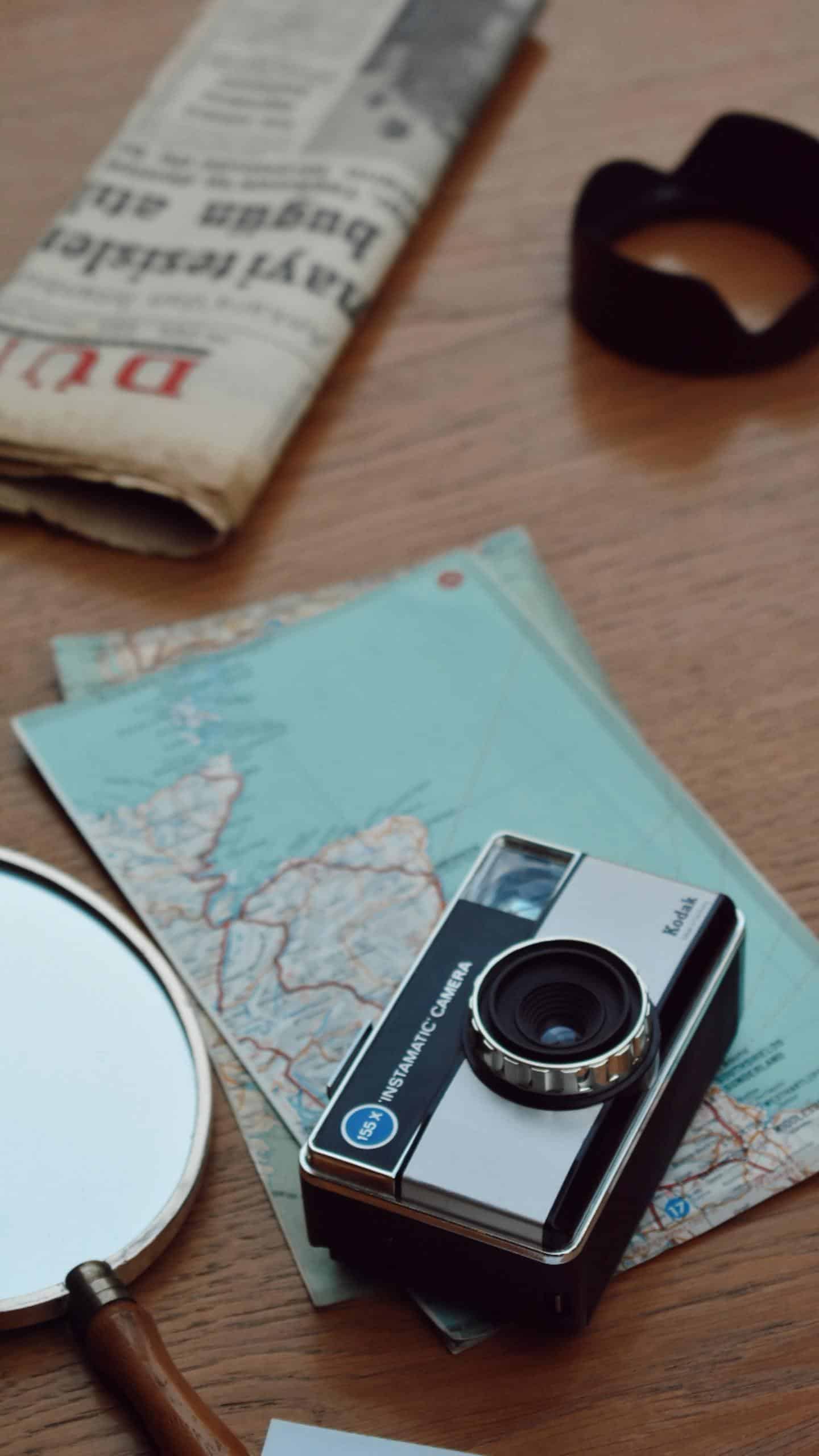 A vintage camera, maps, and newspaper on a wooden table create a nostalgic travel vibe.